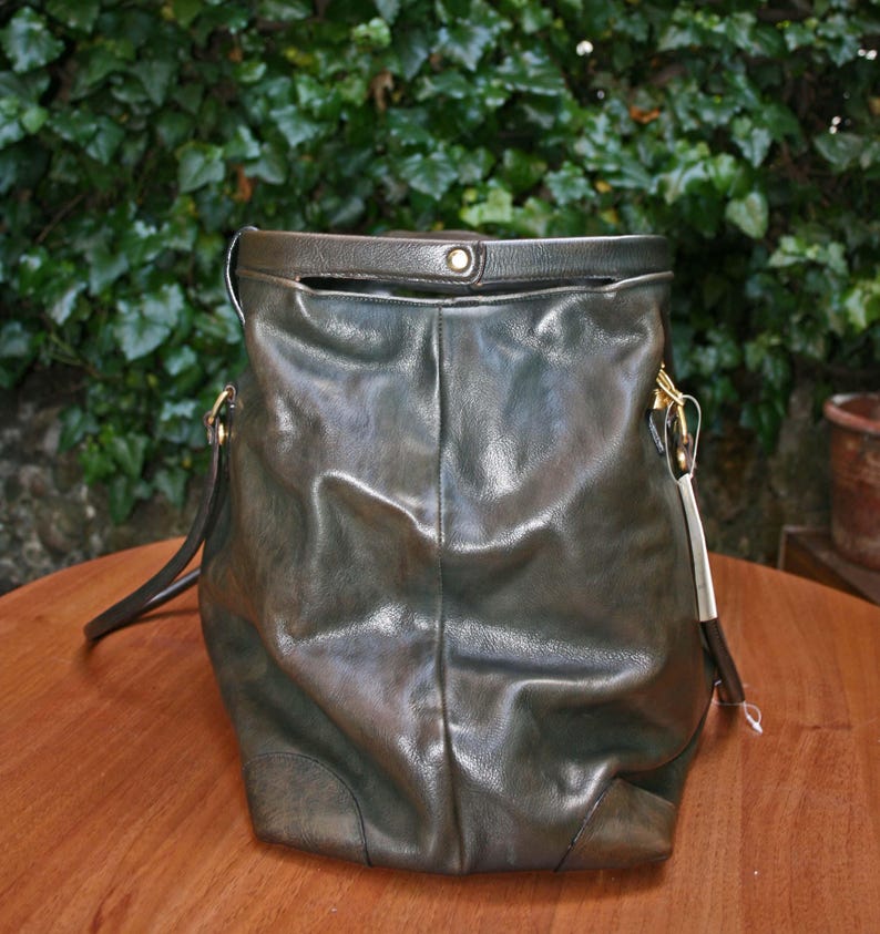 May include: A dark green leather handbag with a shoulder strap and gold-colored hardware. The bag has a soft, slouchy design and a top handle. The background features green foliage and a wooden surface.