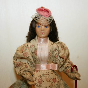 May include: A porcelain doll wearing a pink and white hat with a veil, a floral print dress, and a pink ribbon around the waist.