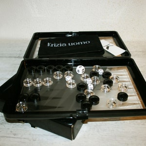 Puede incluir: Una maleta de viaje negra con tapa transparente, que contiene un juego de backgammon con fichas blancas y negras y dados. La maleta tiene una bolsa de tela negra con el texto "Krizia uomo" impreso en ella.