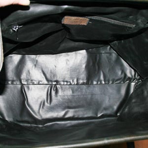 May include: Open view of a black leather bag's interior. The bag has a shiny, smooth lining and a small zippered pocket. Two light-colored straps are visible on the sides. A small brown tag is sewn inside the bag.