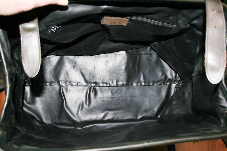 May include: Open view of a black leather bag's interior. The bag has a shiny, smooth lining and a small zippered pocket. Two light-colored straps are visible on the sides. A small brown tag is sewn inside the bag.