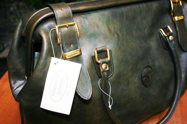 May include: Dark green leather handbag with gold-colored buckles and a tag that reads "valentino orlandi". The bag has a structured shape with a top handle and a shoulder strap. The leather has a slightly distressed appearance.
