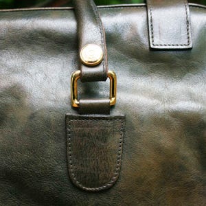 May include: Close-up of a dark green leather handbag. The bag features a gold-colored buckle and a matching gold button on the handle. The leather has a textured appearance.