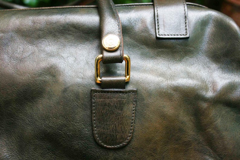 May include: Close-up of a dark green leather handbag. The bag features a gold-colored buckle and a matching gold button on the handle. The leather has a textured appearance.