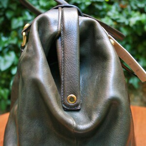 May include: Close-up of a dark olive green leather handbag with a top handle and gold-colored hardware. The bag has a structured design with a slightly worn appearance, suggesting a vintage aesthetic. A tag is attached to the side.