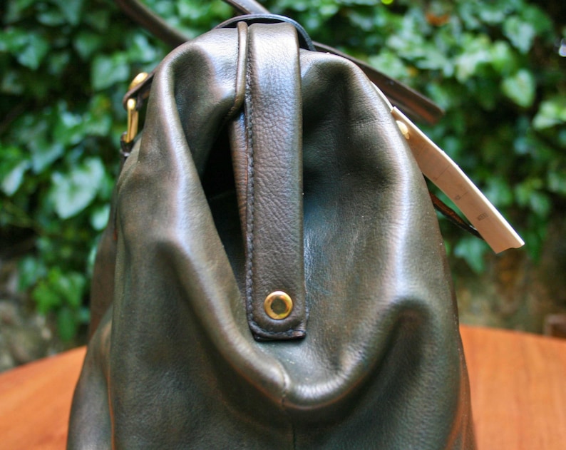 May include: Close-up of a dark olive green leather handbag with a top handle and gold-colored hardware. The bag has a structured design with a slightly worn appearance, suggesting a vintage aesthetic. A tag is attached to the side.