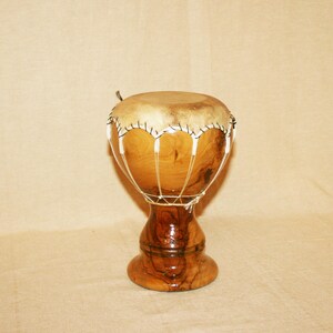 May include: A goblet-shaped drum crafted from polished light brown wood with visible grain patterns. The drum head is made of natural tan animal skin, secured to the wooden body with white cord lacing that creates a decorative scalloped pattern.