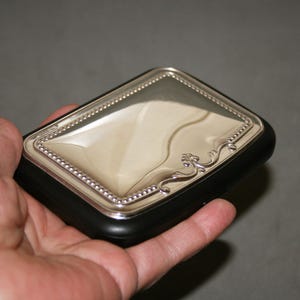May include: A rectangular silver compact with a black base. The silver lid has a beaded border and decorative flourishes. The compact is held in a hand.