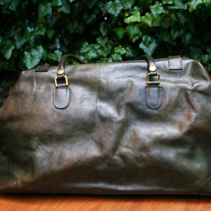 May include: A large, olive-green leather duffel bag with two top handles and gold-colored hardware. The bag has a rectangular shape and appears to be made of distressed leather. The background features green foliage.