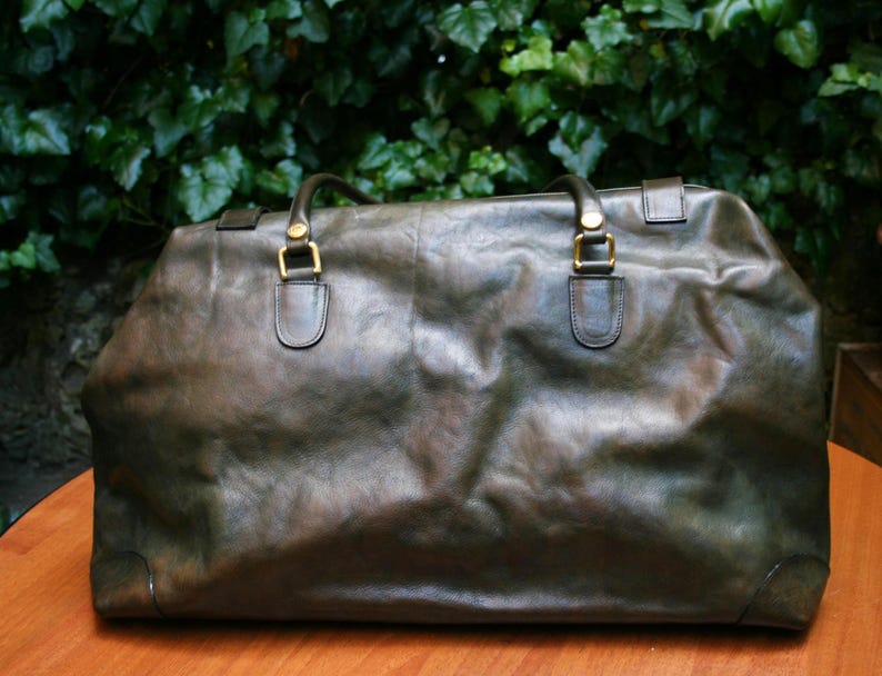 May include: A large, olive-green leather duffel bag with two top handles and gold-colored hardware. The bag has a rectangular shape and appears to be made of distressed leather. The background features green foliage.