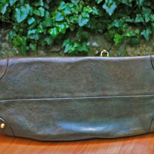 May include: A dark olive green leather handbag with gold-colored corner studs and a matching clasp. The bag has a rectangular shape with a slightly curved top and a textured surface. The bag is sitting on a wooden surface.