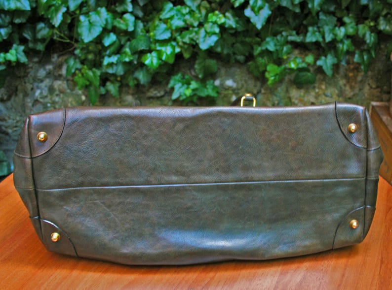 May include: A dark olive green leather handbag with gold-colored corner studs and a matching clasp. The bag has a rectangular shape with a slightly curved top and a textured surface. The bag is sitting on a wooden surface.
