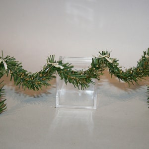 May include: A green garland with gold beads and white bows. The garland is draped over a clear acrylic box.