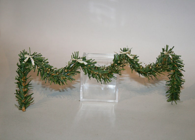 May include: A green garland with gold beads and white bows. The garland is draped over a clear acrylic box.