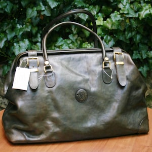 May include: A dark green leather travel bag with a top handle and two shorter handles. The bag has gold-colored buckles and a circular emblem on the front. A white tag hangs from one of the buckles. The bag is sitting on a wooden surface.