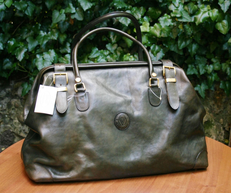 May include: A dark green leather travel bag with a top handle and two shorter handles. The bag has gold-colored buckles and a circular emblem on the front. A white tag hangs from one of the buckles. The bag is sitting on a wooden surface.