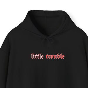 May include: Black hooded sweatshirt with a front pocket and drawstring. The phrase "little trouble" is printed in a red, gothic-style font across the chest. The sweatshirt is made of a soft material.