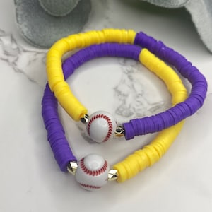 May include: Two bracelets made of colorful, round beads. One bracelet is yellow and the other is purple. Both bracelets have a white baseball charm with red stitching.