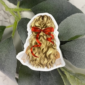 May include: A sticker featuring a woman's blonde hair styled with a red bow. The hair is wavy and detailed, with highlights and shadows. The sticker has a white border and is set against a background of green leaves.