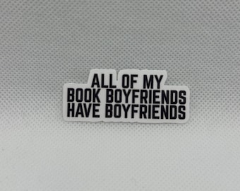 May include: A white sticker with the text "ALL OF MY BOOK BOYFRIENDS HAVE BOYFRIENDS" in black, block letters. The sticker is rectangular with rounded corners and is set against a textured, light gray background.