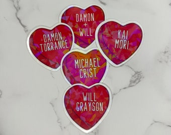 May include: Five holographic heart-shaped stickers with different names and phrases. The stickers are red, pink, and orange with a white outline. The text on the stickers reads: "Damon Torrance", "Damon + Will", "Kai Mori", "Michael Crist", and "Will Grayson".
