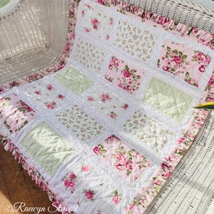 May include: A floral patchwork quilt in pink, white, and green. The quilt has squares of fabric with rose and gingham prints, framed by a ruffled edge. The quilt is on a white wicker chair. The quilt is a charming home decor item.