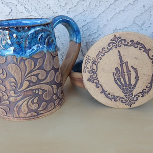 May include: A brown ceramic mug with a blue glaze and a floral pattern. The mug has a handle and is paired with a matching lid featuring a skeleton hand giving the middle finger.