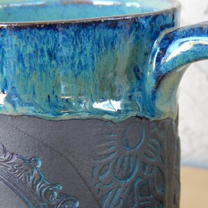 May include: Close-up of a ceramic mug with a blue and turquoise glaze. The mug features a dark gray base with embossed floral and ornate frame designs. The handle is also glazed in blue.