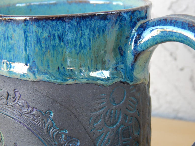May include: Close-up of a ceramic mug with a blue and turquoise glaze. The mug features a dark gray base with embossed floral and ornate frame designs. The handle is also glazed in blue.