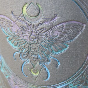 May include: Close-up of a dark gray ceramic mug with an embossed moth design. The moth is detailed with blue, purple, and green accents. The design includes a crescent moon and geometric shapes, creating a gothic or mystical theme.