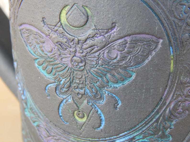 May include: Close-up of a dark gray ceramic mug with an embossed moth design. The moth is detailed with blue, purple, and green accents. The design includes a crescent moon and geometric shapes, creating a gothic or mystical theme.