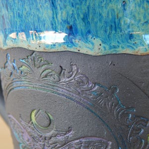 May include: Close-up of a ceramic mug with a detailed, embossed design. The mug features a dark gray base with a blue and purple floral pattern. The top is a vibrant, glossy blue.