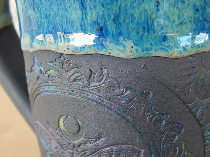 May include: Close-up of a ceramic mug with a detailed, embossed design. The mug features a dark gray base with a blue and purple floral pattern. The top is a vibrant, glossy blue.