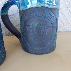 May include: A ceramic mug with a dark blue base and a lighter blue rim. The mug features a detailed embossed design of a moth within a decorative frame. The handle is dark blue. The mug is on a light brown surface.