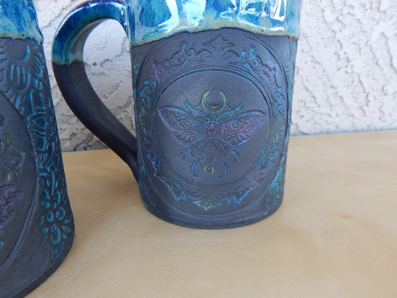 May include: A ceramic mug with a dark blue base and a lighter blue rim. The mug features a detailed embossed design of a moth within a decorative frame. The handle is dark blue. The mug is on a light brown surface.