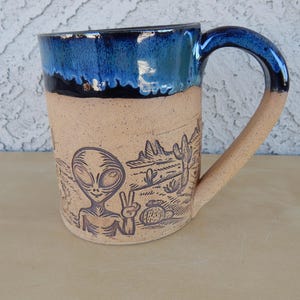 May include: A ceramic mug with a blue glaze and a brown alien with a peace sign on the front. The mug has a desert scene with cacti and mountains in the background.