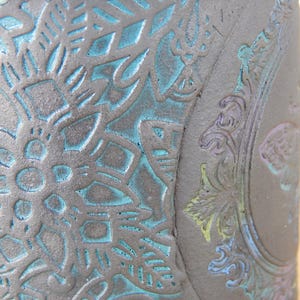 May include: Close-up of a dark gray ceramic vase with intricate embossed floral and ornamental designs. The recessed areas are highlighted with a turquoise-blue color, creating a striking contrast. The vase has a textured surface.