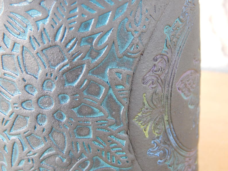 May include: Close-up of a dark gray ceramic vase with intricate embossed floral and ornamental designs. The recessed areas are highlighted with a turquoise-blue color, creating a striking contrast. The vase has a textured surface.
