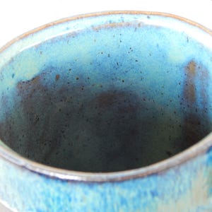 May include: Close-up of a handmade ceramic mug with a blue and turquoise glaze. The interior of the mug has a dark blue hue, and the rim is a lighter shade of blue. The mug has a handle and a dark base.