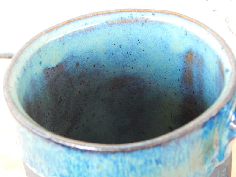 May include: Close-up of a handmade ceramic mug with a blue and turquoise glaze. The interior of the mug has a dark blue hue, and the rim is a lighter shade of blue. The mug has a handle and a dark base.