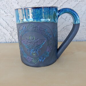 May include: A ceramic mug with a dark gray body and a turquoise blue rim and handle. The mug features an embossed design of a moth within an ornate frame. The handle is a dark gray color. The mug is approximately 4 inches tall.