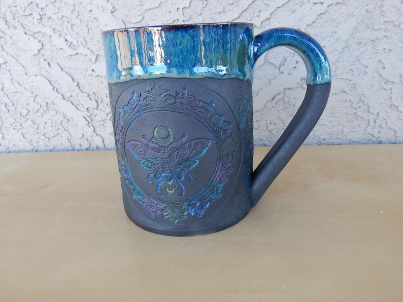 May include: A ceramic mug with a dark gray body and a turquoise blue rim and handle. The mug features an embossed design of a moth within an ornate frame. The handle is a dark gray color. The mug is approximately 4 inches tall.
