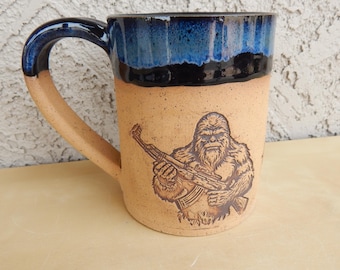 May include: A ceramic mug with a blue and brown glaze. The mug features a brown illustration of a Bigfoot holding a rifle.