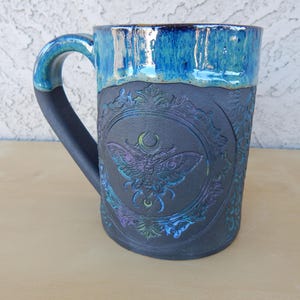 May include: A ceramic mug with a dark gray base and a blue glazed rim. The mug features an embossed design of a moth within a decorative frame. The handle is dark gray, and the mug has a capacity of approximately 16 ounces.