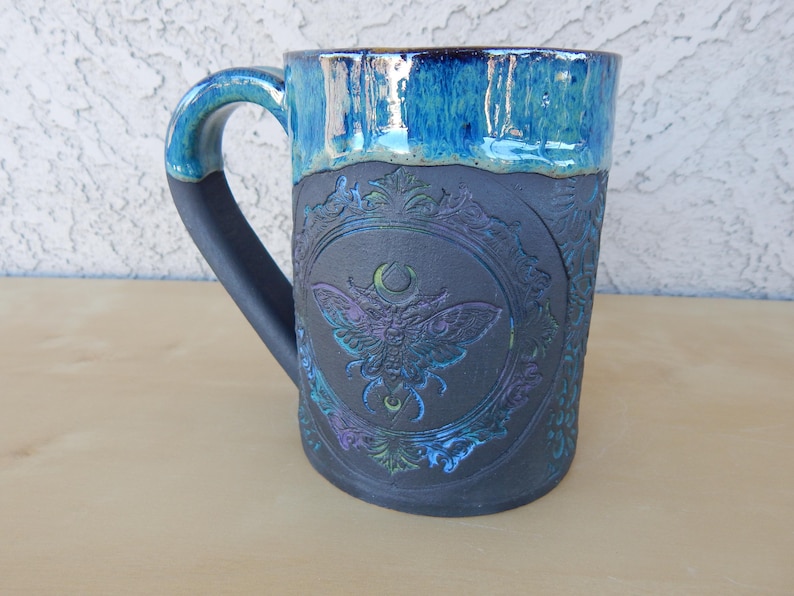 May include: A ceramic mug with a dark gray base and a blue glazed rim. The mug features an embossed design of a moth within a decorative frame. The handle is dark gray, and the mug has a capacity of approximately 16 ounces.