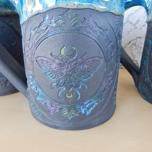 May include: Close-up of three ceramic mugs with a dark gray base and a blue rim. Each mug features a detailed embossed design of a moth within a decorative frame, with iridescent blue and purple accents.