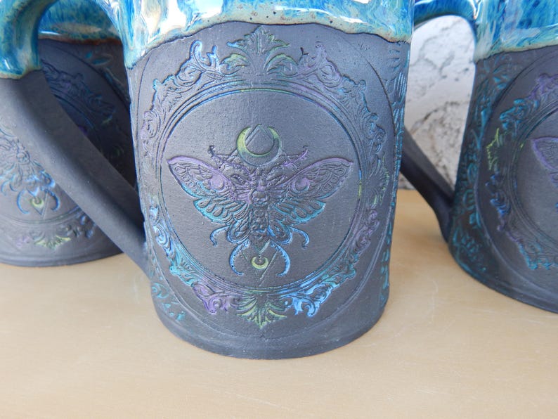 May include: Close-up of three ceramic mugs with a dark gray base and a blue rim. Each mug features a detailed embossed design of a moth within a decorative frame, with iridescent blue and purple accents.