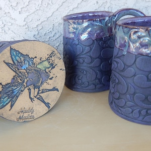 May include: Three ceramic mugs with a purple and blue glaze. The mugs have a raised embossed design of swirling leaves. One mug has a beige bottom with a hand-painted image of a fairy with wings. The bottom of the mug reads "Middy Magian".