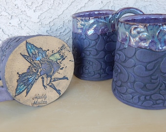 Può includere: Tre tazze in ceramica con una glassa viola e blu. Le tazze hanno un design in rilievo di foglie a spirale. Una tazza ha un fondo beige con un'immagine dipinta a mano di una fata con le ali. Il fondo della tazza recita "Middy Magian".