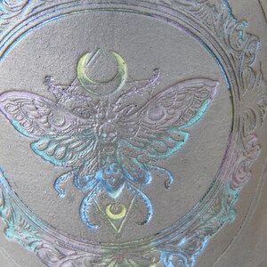 May include: A dark gray ceramic mug with a detailed embossed design. The central image is a moth with a crescent moon and geometric shapes, framed by an ornate border. The moth and border have iridescent blue, green, and purple accents.